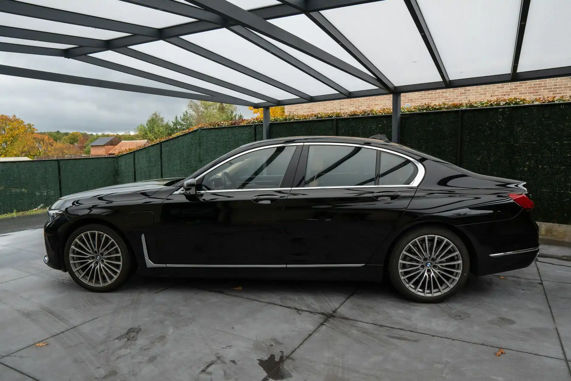black luxury town car pulling up to a covered residential driveway at dawn in Bellevue, Washington, with soft rain reflections on the pavement and packed travel luggage near the...