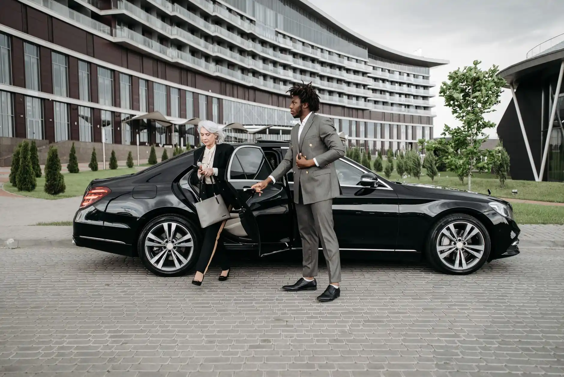professional chauffeur in dark suit standing beside a luxury town car outside a modern Seattle restaurant entrance, city lights reflecting on wet pavement